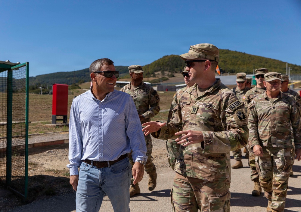 A civilian in casual attire (blue shirt and jeans) is engaged in conversation with a U.S. Army soldier in MultiCam/OCP camouflage uniform while walking outdoors. Several other uniformed military personnel are visible in the background. The setting appears to be a military installation or training area with hills and open terrain visible.