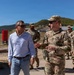 A civilian in casual attire (blue shirt and jeans) is engaged in conversation with a U.S. Army soldier in MultiCam/OCP camouflage uniform while walking outdoors. Several other uniformed military personnel are visible in the background. The setting appears to be a military installation or training area with hills and open terrain visible.