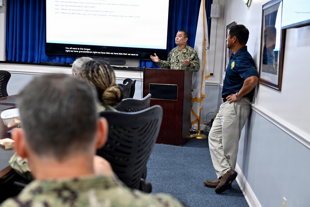 A military briefing or presentation is taking place in a conference room, with a uniformed Navy personnel speaking at a podium in front of a large projection screen. The screen displays text related to health topics, with visible subtitles mentioning living longer and grandchildren. Attendees in military uniforms and civilian attire are seated and standing in the room.