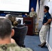 A military briefing or presentation is taking place in a conference room, with a uniformed Navy personnel speaking at a podium in front of a large projection screen. The screen displays text related to health topics, with visible subtitles mentioning living longer and grandchildren. Attendees in military uniforms and civilian attire are seated and standing in the room.
