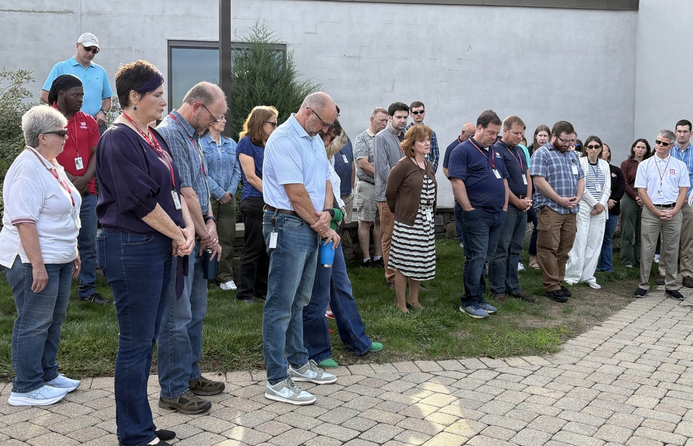 A large group of approximately 25-30 people are gathered outdoors on a brick patio area beside a building, with their heads bowed in what appears to be a moment of silence or prayer. Many individuals are wearing lanyards with ID badges, suggesting this is a workplace or organizational gathering. The solemn posture and unified stance of the group indicates a formal commemorative or reflective event.