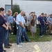 A large group of approximately 25-30 people are gathered outdoors on a brick patio area beside a building, with their heads bowed in what appears to be a moment of silence or prayer. Many individuals are wearing lanyards with ID badges, suggesting this is a workplace or organizational gathering. The solemn posture and unified stance of the group indicates a formal commemorative or reflective event.