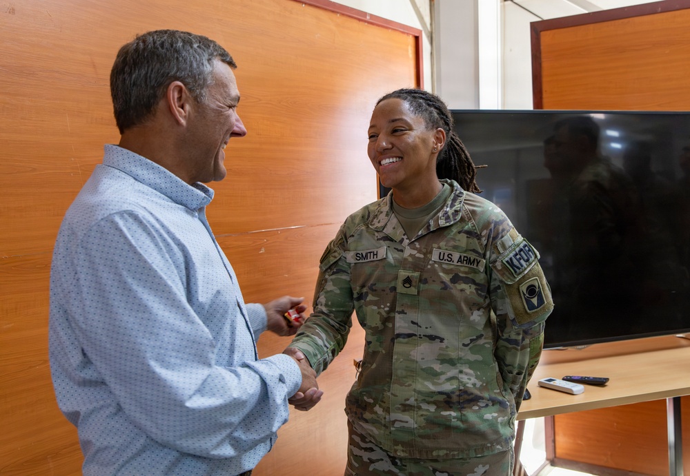 A U.S. Army soldier named Smith, wearing OCP (Operational Camouflage Pattern) uniform with KFOR patch, is engaged in a friendly handshake and conversation with a civilian man in a light blue shirt. Both individuals are smiling warmly, suggesting a positive interaction. The setting appears to be an indoor facility, possibly a military or administrative building.