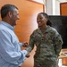 A U.S. Army soldier named Smith, wearing OCP (Operational Camouflage Pattern) uniform with KFOR patch, is engaged in a friendly handshake and conversation with a civilian man in a light blue shirt. Both individuals are smiling warmly, suggesting a positive interaction. The setting appears to be an indoor facility, possibly a military or administrative building.