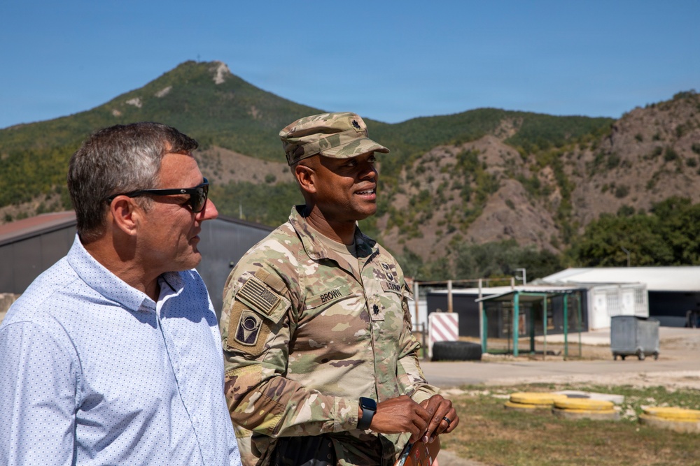 Two individuals stand outdoors at what appears to be a military installation, with a mountainous landscape in the background. One person is wearing a U.S. Army combat uniform with the name tape 'BROWN' and a major's oak leaf insignia on their cap, while the other is in civilian attire. They appear to be engaged in conversation or observing something off-camera.