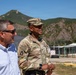Two individuals stand outdoors at what appears to be a military installation, with a mountainous landscape in the background. One person is wearing a U.S. Army combat uniform with the name tape 'BROWN' and a major's oak leaf insignia on their cap, while the other is in civilian attire. They appear to be engaged in conversation or observing something off-camera.