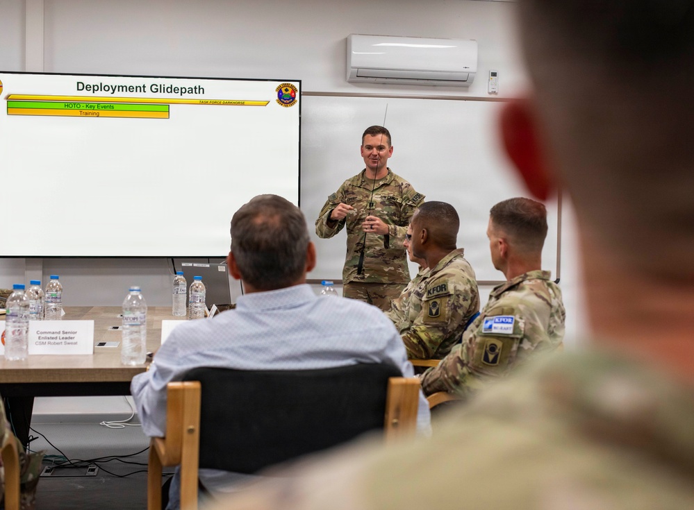 A military briefing is taking place in a conference room where a soldier in combat uniform presents a 'Deployment Glidepath' slide to an audience of military personnel and at least one civilian. The presentation appears to be associated with 'Task Force Darkhorse' and includes a timeline showing 'HOTO - Key Events' and 'Training' phases. Several attendees wearing KFOR (Kosovo Force) patches are visible among the audience.