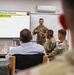A military briefing is taking place in a conference room where a soldier in combat uniform presents a 'Deployment Glidepath' slide to an audience of military personnel and at least one civilian. The presentation appears to be associated with 'Task Force Darkhorse' and includes a timeline showing 'HOTO - Key Events' and 'Training' phases. Several attendees wearing KFOR (Kosovo Force) patches are visible among the audience.