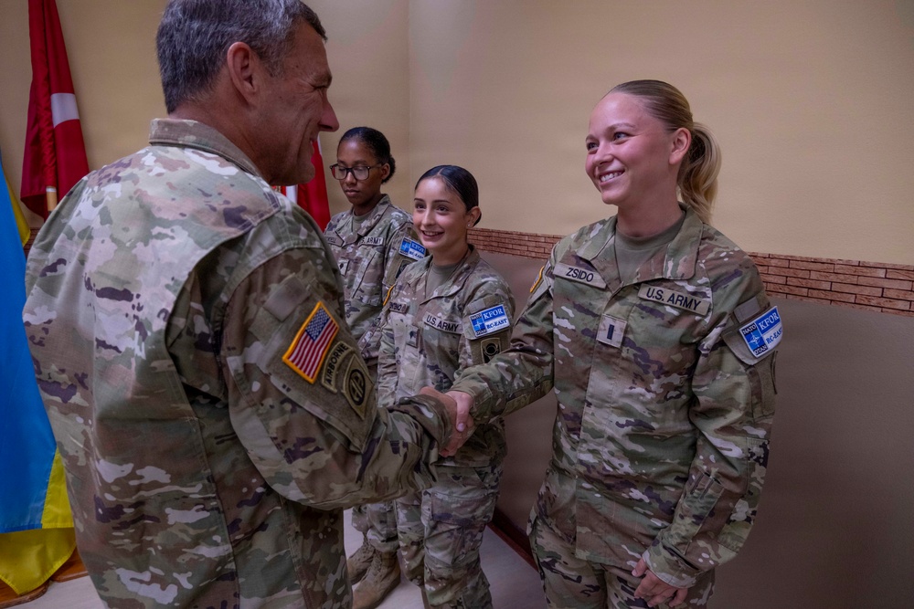 A U.S. Army senior officer is shaking hands with soldiers during what appears to be a formal recognition or greeting ceremony. The soldiers are wearing OCP (Operational Camouflage Pattern) uniforms with KFOR (Kosovo Force) NATO patches, indicating deployment to Kosovo. Three female soldiers are visible, with one named 'ZSIDO' prominently in the foreground.