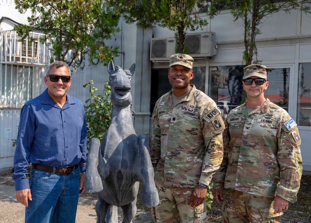 Three individuals pose for a photo outdoors near a stone or metal horse statue. Two of the individuals are wearing U.S. Army combat uniforms with KFOR (Kosovo Force) patches, while the third is in civilian clothing. The setting appears to be a military installation or compound.