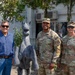 Three individuals pose for a photo outdoors near a stone or metal horse statue. Two of the individuals are wearing U.S. Army combat uniforms with KFOR (Kosovo Force) patches, while the third is in civilian clothing. The setting appears to be a military installation or compound.