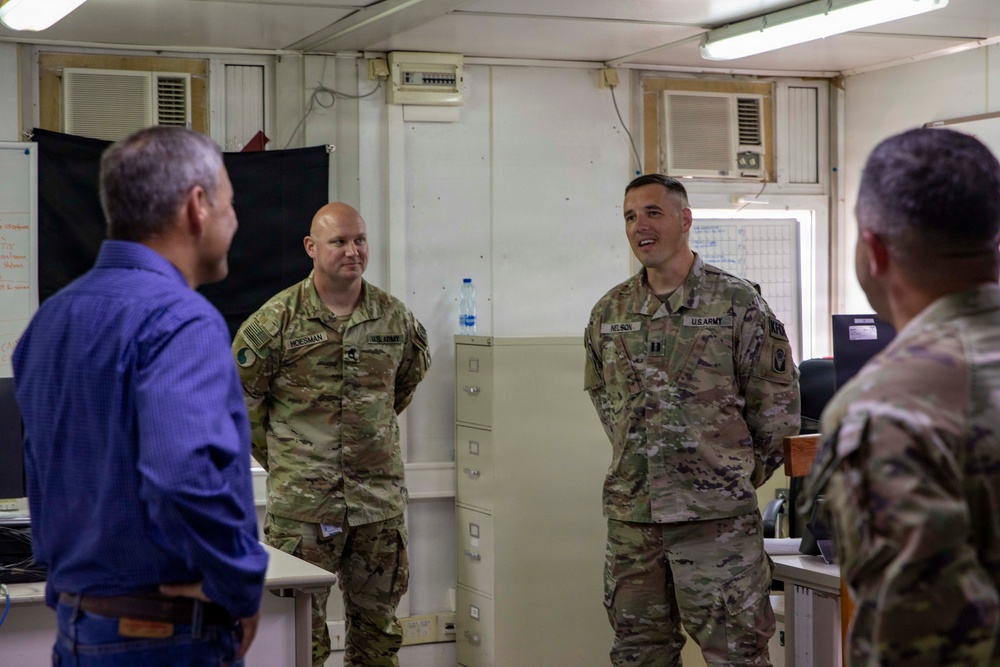Four individuals are gathered in what appears to be a military operations or administrative room. Two U.S. Army soldiers in OCP (Operational Camouflage Pattern) uniforms, identified by name tapes as 'HOESMAN' and 'NELSON', are conversing with a civilian in a blue shirt and another military personnel whose back is to the camera. The setting appears to be a forward operating base or military installation office.