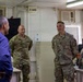 Four individuals are gathered in what appears to be a military operations or administrative room. Two U.S. Army soldiers in OCP (Operational Camouflage Pattern) uniforms, identified by name tapes as 'HOESMAN' and 'NELSON', are conversing with a civilian in a blue shirt and another military personnel whose back is to the camera. The setting appears to be a forward operating base or military installation office.