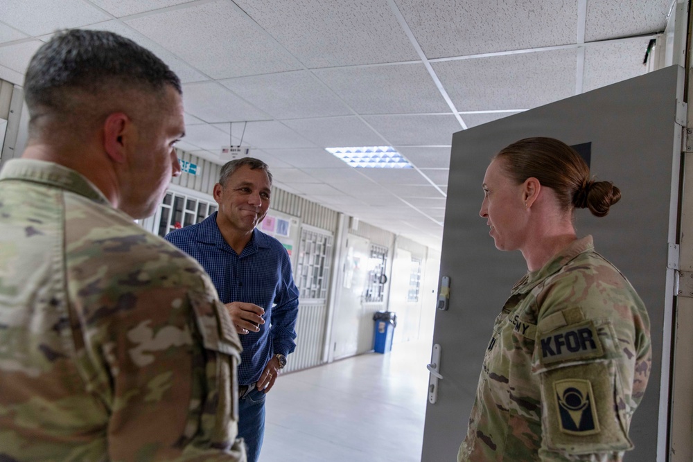 Three individuals are engaged in conversation in what appears to be a military facility corridor. Two are wearing U.S. Army OCP (Operational Camouflage Pattern) uniforms with KFOR (Kosovo Force) patches, while one civilian male in a blue shirt participates in the discussion. The setting suggests an official visit or briefing at a NATO KFOR installation.