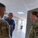 Three individuals are engaged in conversation in what appears to be a military facility corridor. Two are wearing U.S. Army OCP (Operational Camouflage Pattern) uniforms with KFOR (Kosovo Force) patches, while one civilian male in a blue shirt participates in the discussion. The setting suggests an official visit or briefing at a NATO KFOR installation.