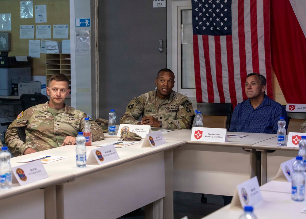 Three individuals are seated at a conference table in what appears to be a military briefing room. Two are wearing U.S. Army combat uniforms (ACUs) and one is in civilian attire. An American flag and what appears to be a Polish flag are visible in the background.