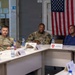 Three individuals are seated at a conference table in what appears to be a military briefing room. Two are wearing U.S. Army combat uniforms (ACUs) and one is in civilian attire. An American flag and what appears to be a Polish flag are visible in the background.
