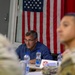 A middle-aged civilian man wearing glasses and a blue plaid shirt sits at a conference table in front of a displayed American flag. He appears to be attending a military briefing or meeting, with uniformed military personnel visible in the foreground (out of focus). Water bottles, documents, and a placard with a red shield insignia are visible on the table.