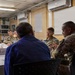 A military briefing or meeting is taking place in what appears to be a forward operating base or military facility. Several personnel in U.S. Army OCP (Operational Camouflage Pattern) uniforms are seated at a table alongside a civilian in a blue checkered shirt. A Florida state flag is visible in the background, and name placards are present on the table.