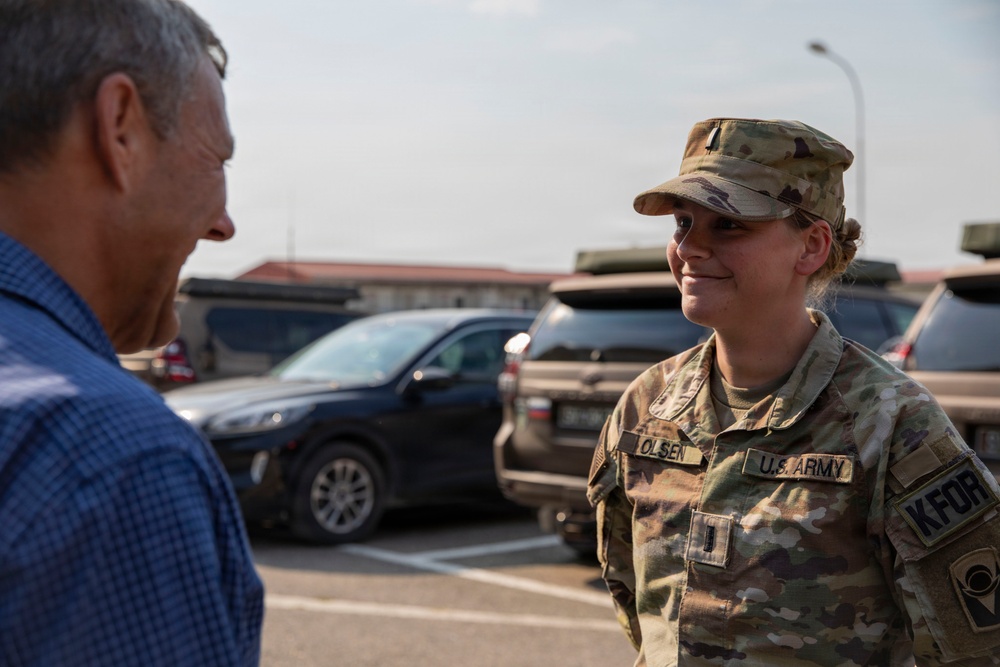 A U.S. Army soldier identified by name tape as 'OLSEN' is engaged in conversation with a civilian male in a parking lot. The soldier is wearing Operational Camouflage Pattern (OCP) uniform with a KFOR (Kosovo Force) patch visible on the right shoulder, indicating deployment to Kosovo. The interaction appears cordial and informal.