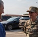 A U.S. Army soldier identified by name tape as 'OLSEN' is engaged in conversation with a civilian male in a parking lot. The soldier is wearing Operational Camouflage Pattern (OCP) uniform with a KFOR (Kosovo Force) patch visible on the right shoulder, indicating deployment to Kosovo. The interaction appears cordial and informal.