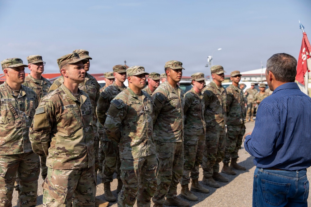 A civilian in a blue plaid shirt and jeans addresses a formation of U.S. Army soldiers in OCP (Operational Camouflage Pattern) uniforms outdoors. The soldiers are standing at attention or at ease in organized rows, suggesting a formal briefing or inspection event. A red military guidon flag is visible in the background, indicating a unit formation.