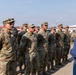 A civilian in a blue plaid shirt and jeans addresses a formation of U.S. Army soldiers in OCP (Operational Camouflage Pattern) uniforms outdoors. The soldiers are standing at attention or at ease in organized rows, suggesting a formal briefing or inspection event. A red military guidon flag is visible in the background, indicating a unit formation.