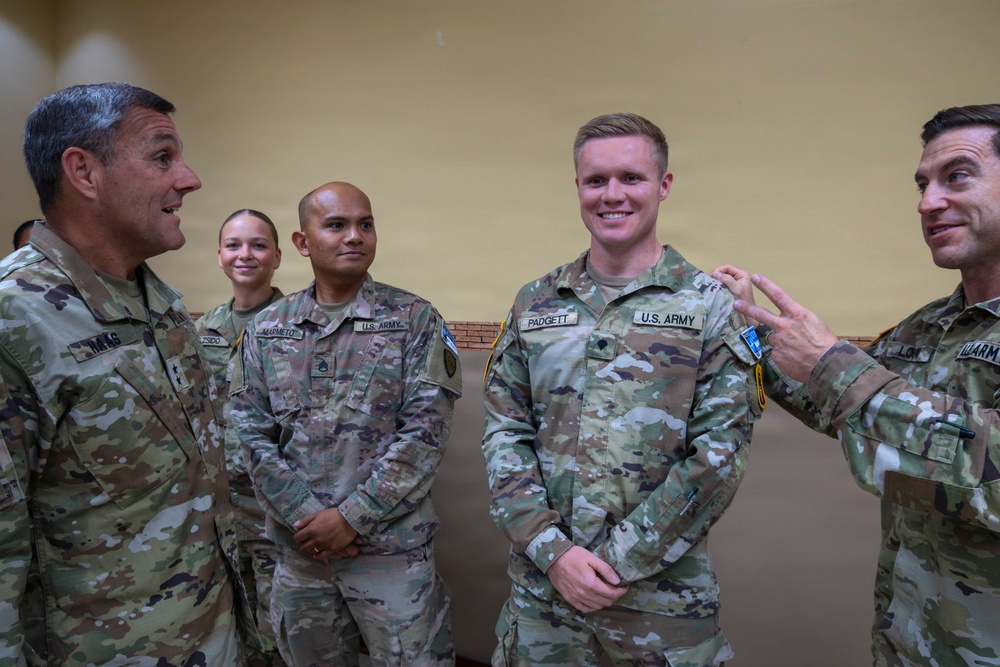 A group of U.S. Army soldiers in OCP (Operational Camouflage Pattern) uniforms are engaged in a casual conversation, appearing relaxed and smiling. The central soldier, identified by the name tape 'PADGETT,' is being addressed by a soldier on the right while another senior soldier on the left also participates. The setting appears to be an indoor military facility with a plain yellow wall in the background.