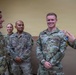 A group of U.S. Army soldiers in OCP (Operational Camouflage Pattern) uniforms are engaged in a casual conversation, appearing relaxed and smiling. The central soldier, identified by the name tape 'PADGETT,' is being addressed by a soldier on the right while another senior soldier on the left also participates. The setting appears to be an indoor military facility with a plain yellow wall in the background.