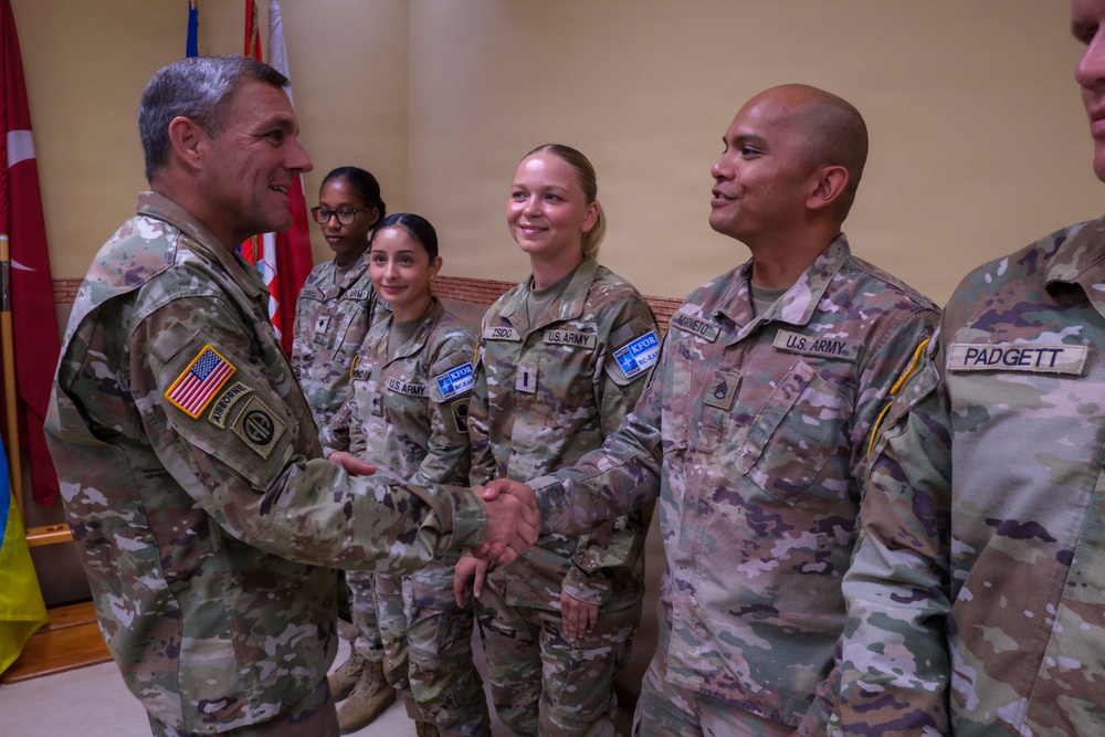 A senior U.S. Army officer wearing an Airborne division patch is shaking hands with a soldier identified by nametape as 'MAMETO' during what appears to be a formal military ceremony or recognition event. Several other U.S. Army soldiers are present in the background, all wearing Operational Camouflage Pattern (OCP) uniforms with KFOR (Kosovo Force) NATO patches. The setting appears to be an indoor military facility with flags visible in the background.