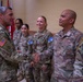 A senior U.S. Army officer wearing an Airborne division patch is shaking hands with a soldier identified by nametape as 'MAMETO' during what appears to be a formal military ceremony or recognition event. Several other U.S. Army soldiers are present in the background, all wearing Operational Camouflage Pattern (OCP) uniforms with KFOR (Kosovo Force) NATO patches. The setting appears to be an indoor military facility with flags visible in the background.