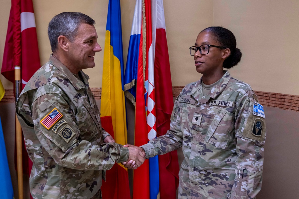 Two U.S. Army soldiers in OCP (Operational Camouflage Pattern) uniforms are shaking hands in what appears to be an official military ceremony or recognition event. The male soldier on the left wears 82nd Airborne Division patches and a U.S. flag patch, while the female soldier on the right has a name tape reading 'DOWDELL' and a U.S. Army tape. Several international flags are displayed in the background.