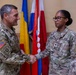 Two U.S. Army soldiers in OCP (Operational Camouflage Pattern) uniforms are shaking hands in what appears to be an official military ceremony or recognition event. The male soldier on the left wears 82nd Airborne Division patches and a U.S. flag patch, while the female soldier on the right has a name tape reading 'DOWDELL' and a U.S. Army tape. Several international flags are displayed in the background.