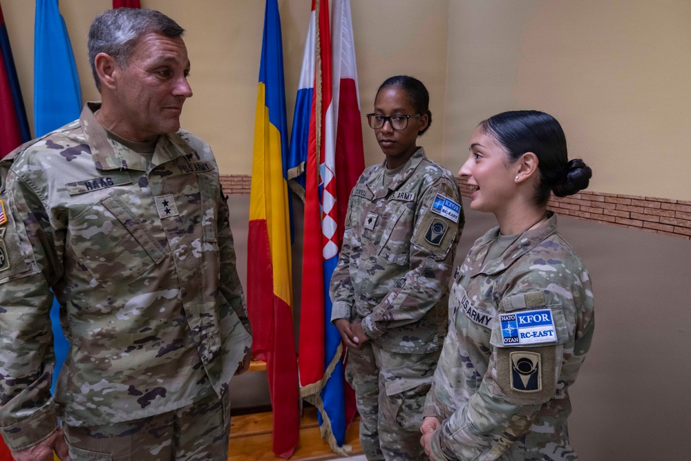 Three U.S. Army soldiers in OCP (Operational Camouflage Pattern) uniforms are engaged in conversation inside a building with multiple national flags in the background. The soldier on the left displays two stars on his chest indicating the rank of Major General, with a name tape reading 'RAAS'. The two soldiers on the right wear KFOR RC-EAST (Kosovo Force Regional Command East) patches, indicating a NATO peacekeeping mission context.