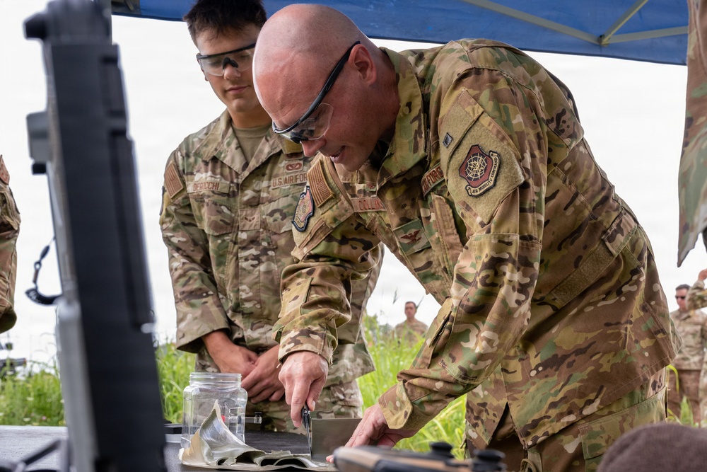 Two U.S. Air Force personnel in OCP (Operational Camouflage Pattern) uniforms are working at an outdoor field station under a blue canopy. The foreground individual, identified by nametape as 'COLLINS,' is leaning over a table performing a hands-on task with a tool, while the second individual ('DEPUCCHI') observes. Both are wearing protective eyewear, suggesting a safety-sensitive activity.