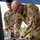 Two U.S. Air Force personnel in OCP (Operational Camouflage Pattern) uniforms are working at an outdoor field station under a blue canopy. The foreground individual, identified by nametape as 'COLLINS,' is leaning over a table performing a hands-on task with a tool, while the second individual ('DEPUCCHI') observes. Both are wearing protective eyewear, suggesting a safety-sensitive activity.