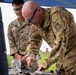 Two U.S. Air Force personnel in OCP (Operational Camouflage Pattern) uniforms are working at an outdoor field station under a blue canopy. The foreground individual, identified by nametape as 'COLLINS,' is leaning over a table performing a hands-on task with a tool, while the second individual ('DEPUCCHI') observes. Both are wearing protective eyewear, suggesting a safety-sensitive activity.