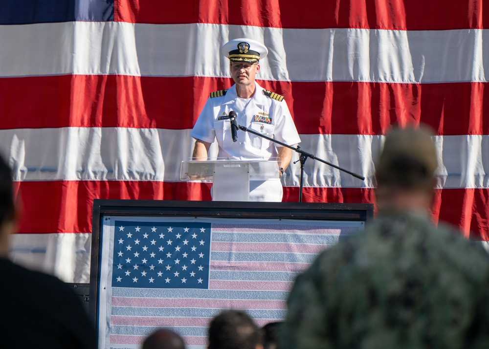 A U.S. Navy officer in white dress uniform stands at a clear podium delivering a speech at a formal military ceremony. A large American flag serves as a backdrop, and a framed display featuring an American flag with text is mounted on the front of the podium. An audience including military personnel in camouflage uniforms is visible in the foreground.