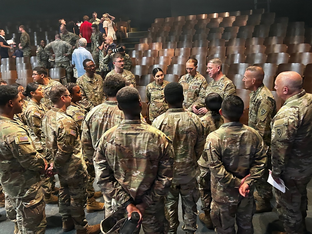 A large group of U.S. Army soldiers in OCP (Operational Camouflage Pattern) uniforms are gathered in what appears to be an auditorium or theater setting. Senior officers appear to be addressing or interacting with junior enlisted personnel and lower-ranking soldiers in a semi-circular formation. The setting suggests a post-event gathering, briefing, or meet-and-greet following a formal ceremony or presentation.