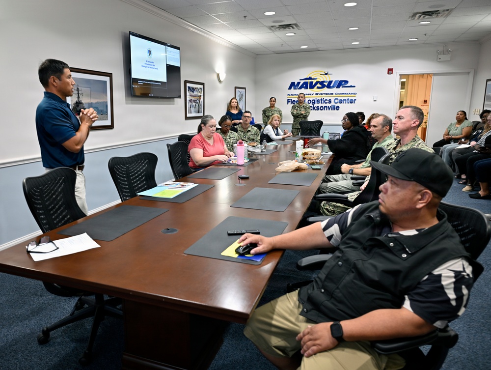 A briefing or meeting is taking place at NAVSUP Fleet Logistics Center Jacksonville, as indicated by the signage on the wall. A presenter stands at the head of a conference table addressing a mixed group of military personnel in Navy uniforms and civilians. A presentation is displayed on a wall-mounted monitor in the background.