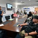 A briefing or meeting is taking place at NAVSUP Fleet Logistics Center Jacksonville, as indicated by the signage on the wall. A presenter stands at the head of a conference table addressing a mixed group of military personnel in Navy uniforms and civilians. A presentation is displayed on a wall-mounted monitor in the background.