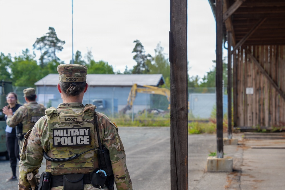 A U.S. Army Military Police soldier with the name tape 'MOODY' is seen from behind, wearing OCP (Operational Camouflage Pattern) uniform and a tactical vest marked 'MILITARY POLICE'. The soldier appears to be on duty at what looks like a military installation or training area, with other personnel visible in the background. A yellow excavator or construction equipment is visible in the mid-ground near a fenced area.