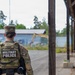 A U.S. Army Military Police soldier with the name tape 'MOODY' is seen from behind, wearing OCP (Operational Camouflage Pattern) uniform and a tactical vest marked 'MILITARY POLICE'. The soldier appears to be on duty at what looks like a military installation or training area, with other personnel visible in the background. A yellow excavator or construction equipment is visible in the mid-ground near a fenced area.