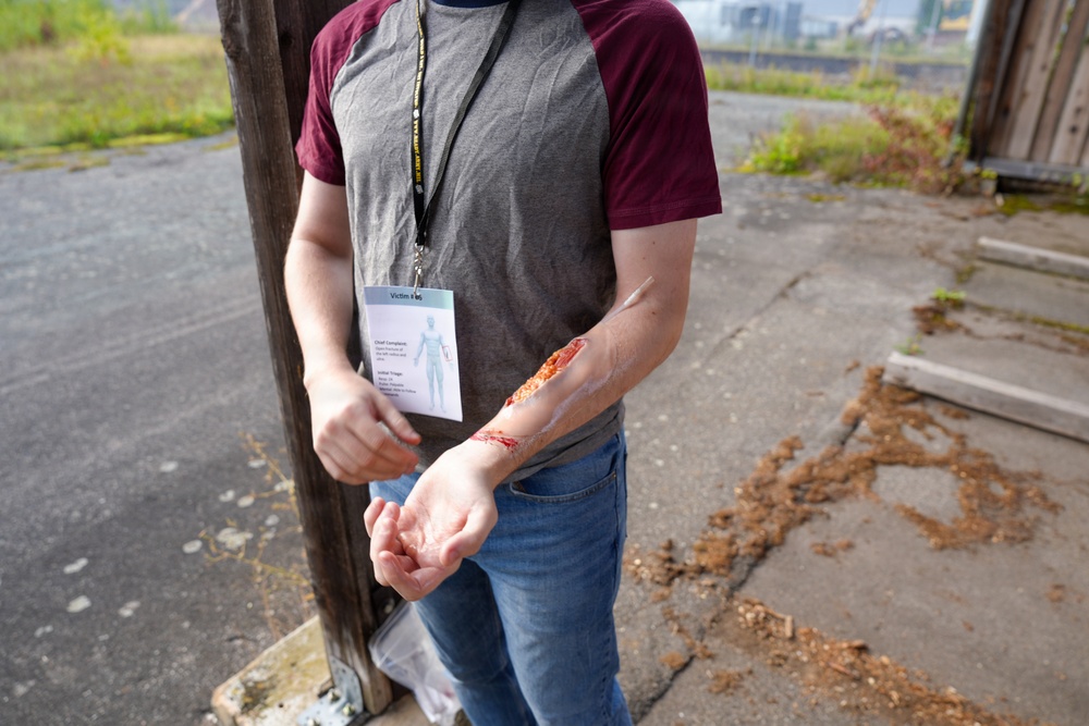 A person wearing a lanyard badge labeled 'Victim' is displaying a simulated wound on their forearm, consistent with a mass casualty exercise or emergency response training drill. The wound appears to be theatrical makeup or moulage applied to simulate a laceration or abrasion injury. The outdoor industrial setting and victim role card suggest this is a structured emergency preparedness simulation.