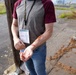 A person wearing a lanyard badge labeled 'Victim' is displaying a simulated wound on their forearm, consistent with a mass casualty exercise or emergency response training drill. The wound appears to be theatrical makeup or moulage applied to simulate a laceration or abrasion injury. The outdoor industrial setting and victim role card suggest this is a structured emergency preparedness simulation.