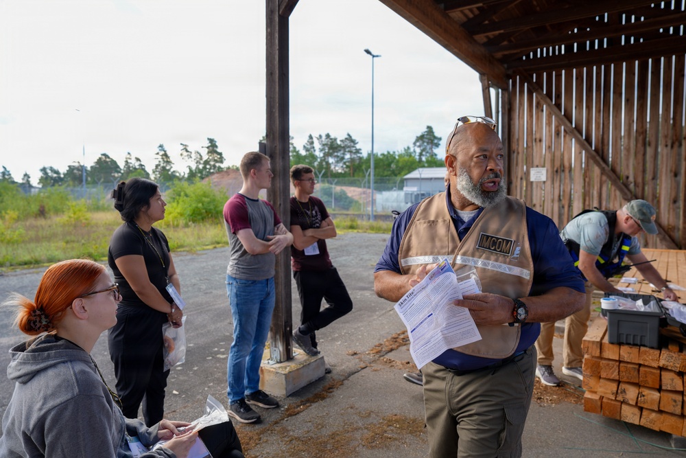 A man wearing an 'IMCOM' vest is addressing or briefing a small group of people under a wooden shelter structure in an outdoor setting. He is holding papers and appears to be speaking, while several individuals stand or sit nearby listening. The setting appears to be a training, inspection, or operational briefing site.