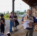 A man wearing an 'IMCOM' vest is addressing or briefing a small group of people under a wooden shelter structure in an outdoor setting. He is holding papers and appears to be speaking, while several individuals stand or sit nearby listening. The setting appears to be a training, inspection, or operational briefing site.