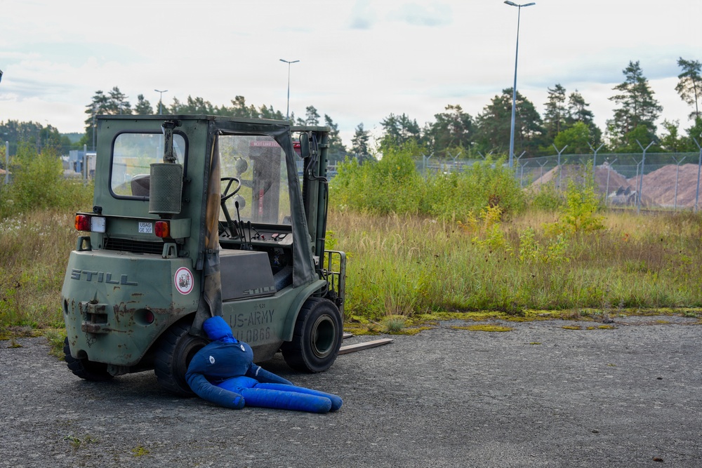 A military-marked 'STILL' brand forklift painted in olive green with 'US-ARMY' markings is parked on a paved surface near an overgrown field. A blue human-shaped dummy or mannequin is positioned on the ground beneath the front forks of the forklift, suggesting a safety training or accident simulation scenario. The setting appears to be an industrial or military facility with fencing, trees, and street lights visible in the background.