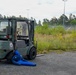 A military-marked 'STILL' brand forklift painted in olive green with 'US-ARMY' markings is parked on a paved surface near an overgrown field. A blue human-shaped dummy or mannequin is positioned on the ground beneath the front forks of the forklift, suggesting a safety training or accident simulation scenario. The setting appears to be an industrial or military facility with fencing, trees, and street lights visible in the background.