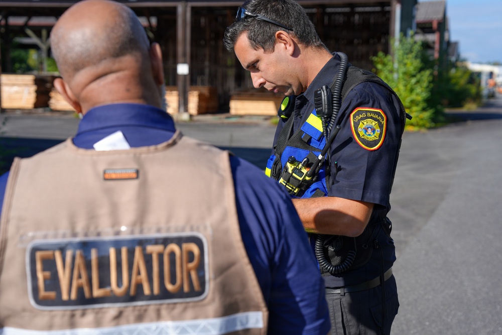 Two individuals are engaged in what appears to be a training exercise or evaluation scenario outdoors. One person is wearing a tan vest labeled 'EVALUATOR' on the back, while the other is a uniformed responder wearing a 'USAG Bavaria' patch and tactical gear including a radio. The setting appears to be an industrial or storage yard with stacked lumber visible in the background.